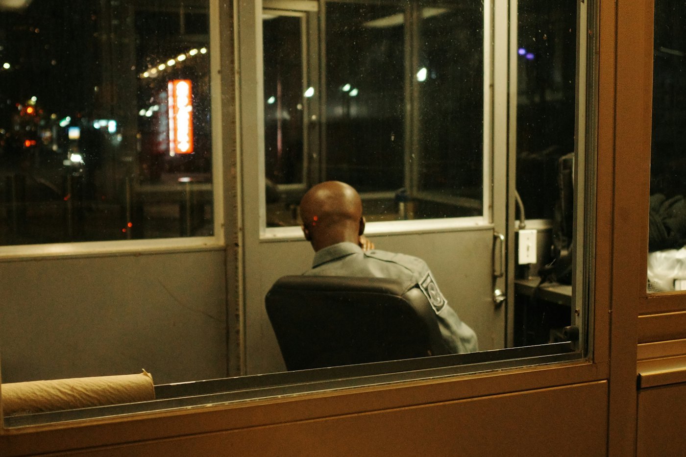 A person sitting alone in a lit security booth at night.